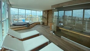 A modern lounge area with large windows, a circular seating area, and a view of the cityscape. - Edifício Promenade II Apt 306 (Maceió)