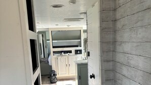 Un couloir étroit doté d’un plancher en bois, d’un téléviseur encastré et d’un coin cuisine avec des placards et un évier.