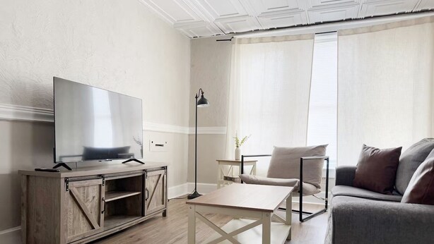 Ein modernes Wohnzimmer mit Fernseher auf einem Holzschrank, einem weißen Couchtisch, einem grauen Sofa und einer Stehlampe.