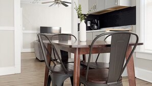 Ein Essbereich mit Holztisch und Metallstühlen, einem Deckenventilator sowie einer Küche mit weißen Schränken und einer schwarzen Spritzschutzwand.