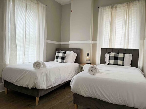 Two single beds with white linens, a small bedside table with a lamp, and sheer curtains. - Cherry Street Retreat A (Evansville)