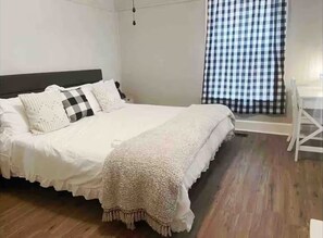 A bedroom with a bed, pillows, a blanket, a wooden floor, and a checkered curtain. - Cherry Street Retreat A (Evansville)