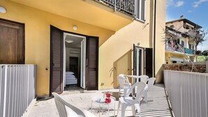 Une terrasse extérieure ensoleillée, avec des chaises en plastique blanches et une petite table, un bâtiment jaune aux volets ouverts et un balcon doté d’une balustrade.