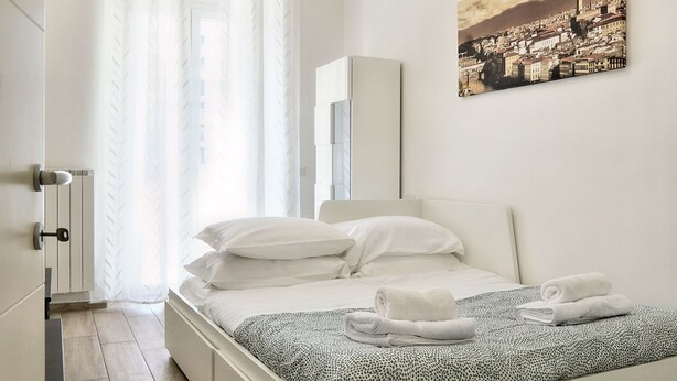 Une chambre à coucher avec un lit, des rideaux blancs, un radiateur et un tableau représentant un paysage urbain accroché au mur.