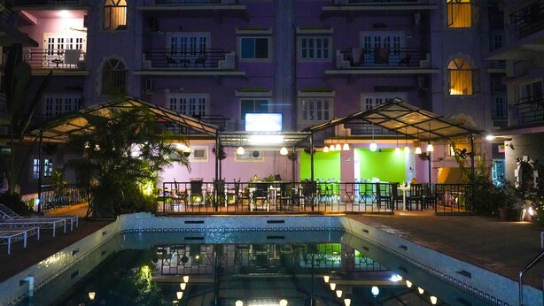 Un área de piscina con una zona de estar techada, una pared verde y un edificio con varios balcones al fondo.