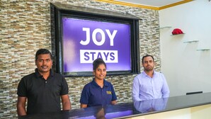 Tres personas de pie en un mostrador de recepción, con un letrero que dice 'JOY STAYS' al fondo.