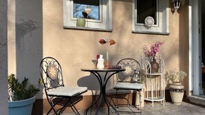 Ein Sitzbereich im Freien mit zwei Metallstühlen und einem kleinen runden Tisch, umgeben von Topfpflanzen und einer dekorativen Vase mit Blumen.