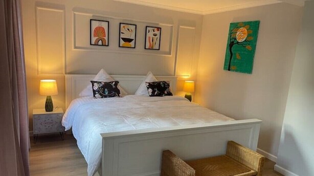 Ein Schlafzimmer mit einem großen Bett, zwei Nachttischlampen, einem Korbstuhl und drei gerahmten Kunstwerken an der Wand.