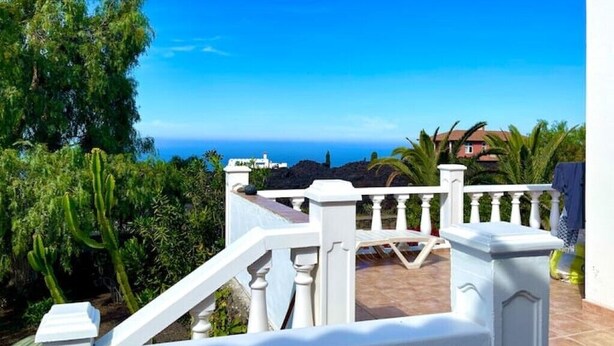 Een balkon met een witte balustrade, met uitzicht op een kustlandschap met groen en een helderblauwe lucht.