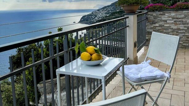 Un balcón con mesa, dos sillas, un cuenco de limones y vistas al mar y a las montañas.