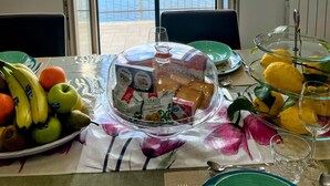 Una mesa de comedor con vistas al mar a través de un ventanal grande, un frutero y un vaso de agua.