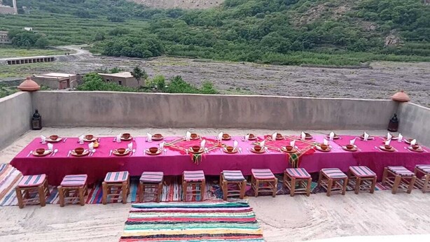 A long table set for a meal with chairs, a colorful rug, and a view of a green landscape. - Riad Dar Tagine (Asni)