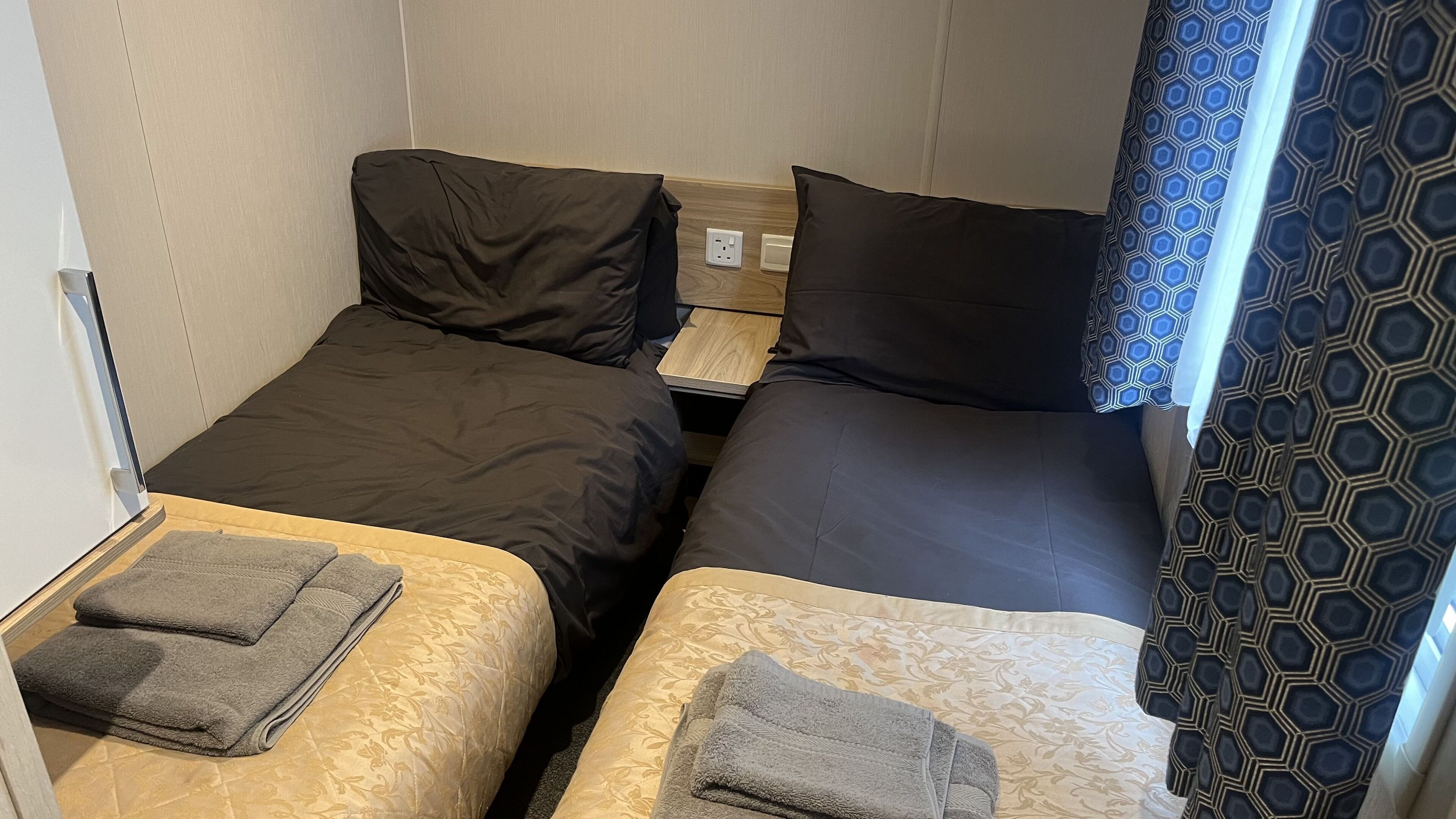 Ein kleiner, kompakter Raum mit zwei Einzelbetten, jeweils mit Kissen und einem Handtuch. Dazwischen befindet sich ein Nachttisch aus Holz, und an der Wand ist eine Steckdose sichtbar.