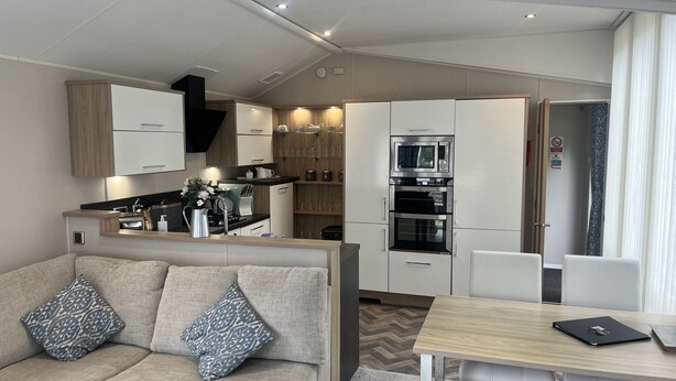 A modern kitchenette with built-in appliances, a dining area with a table and chairs, and a sofa with patterned cushions. - Cosy 3-bed hot tub Lodge in Pr3 (Preston)