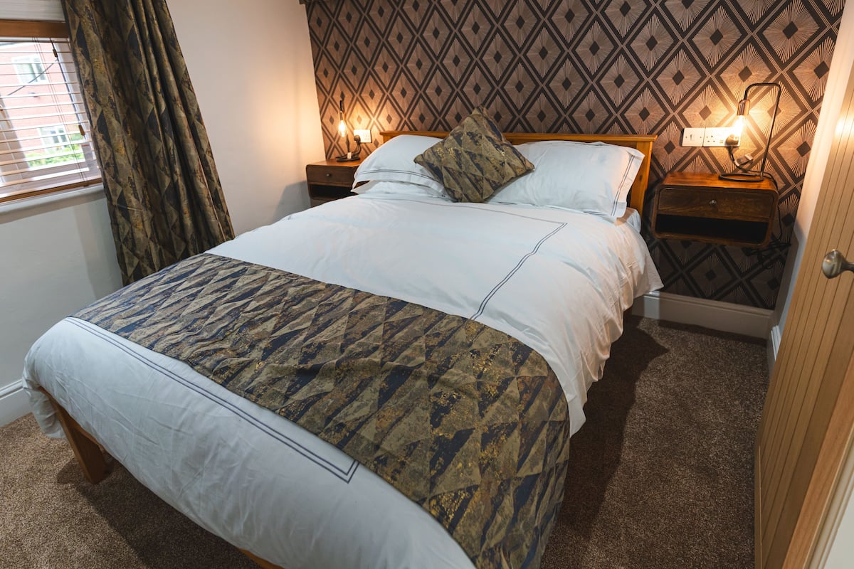A neatly made bed with a patterned bedspread, two bedside tables with lamps, a patterned wallpaper, and a window with curtains.