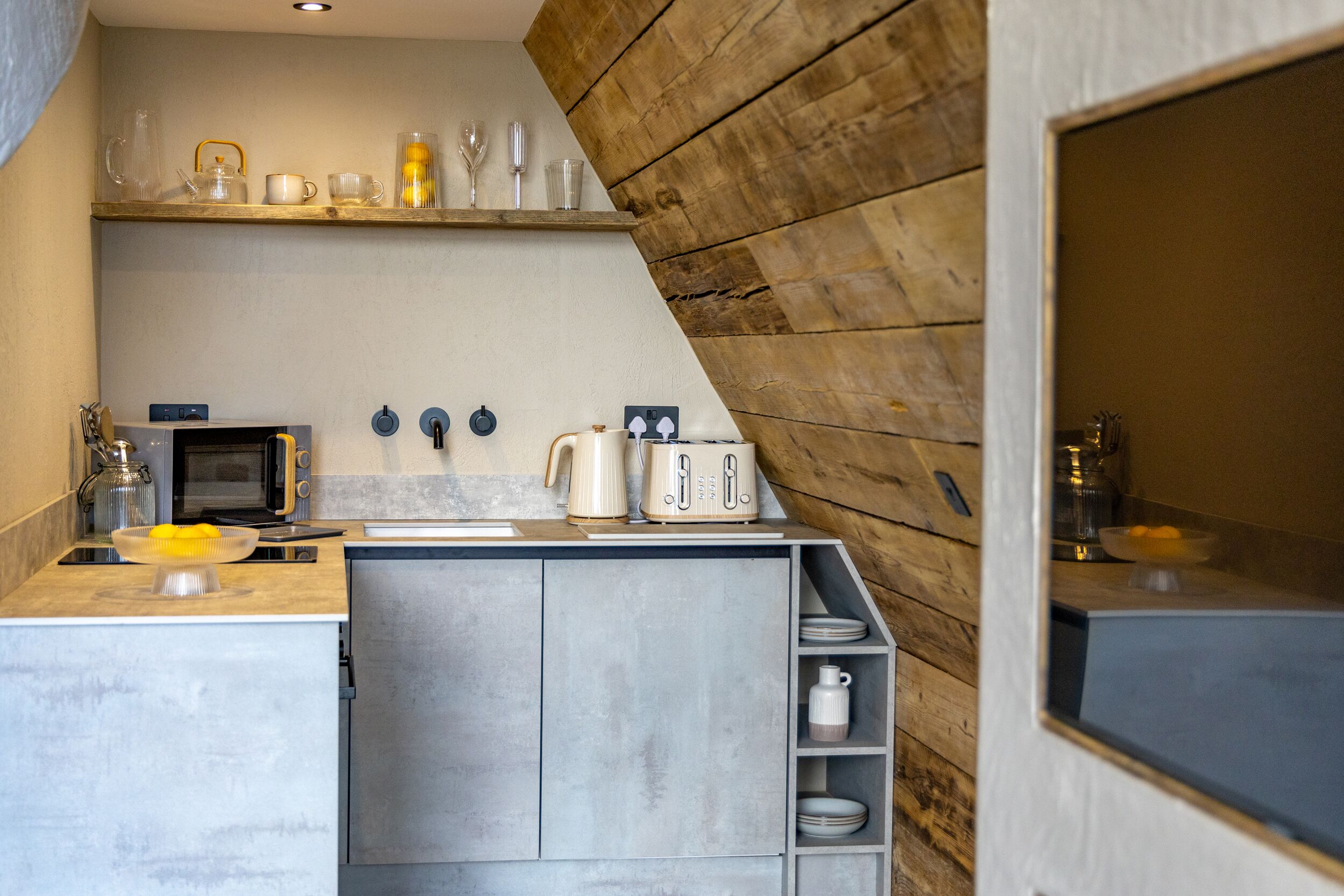 Una cocina moderna con horno empotrado, microondas y tostadora. Hay estantes sobre la encimera con diversos utensilios de cocina y una ventana a la derecha.