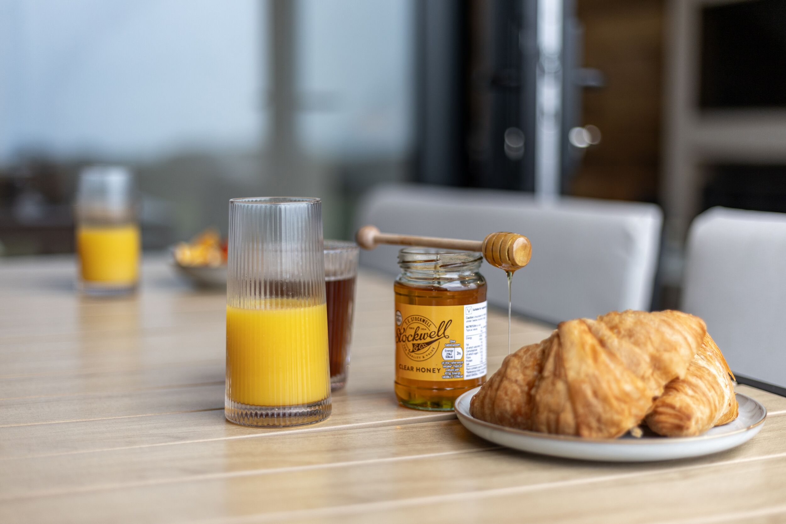 A breakfast setting with a glass of orange juice, a jar of honey, and a croissant on a table.