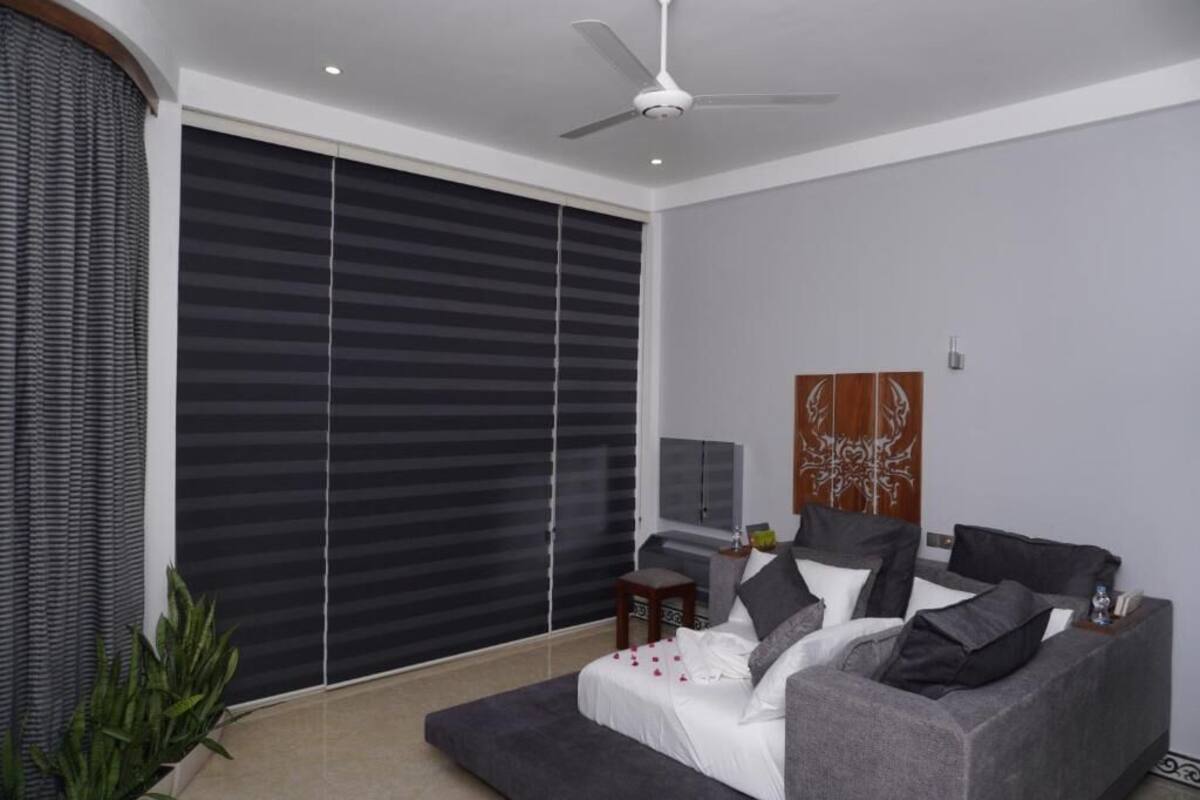 A modern living room with a grey sofa, a ceiling fan, and a large window with blinds.