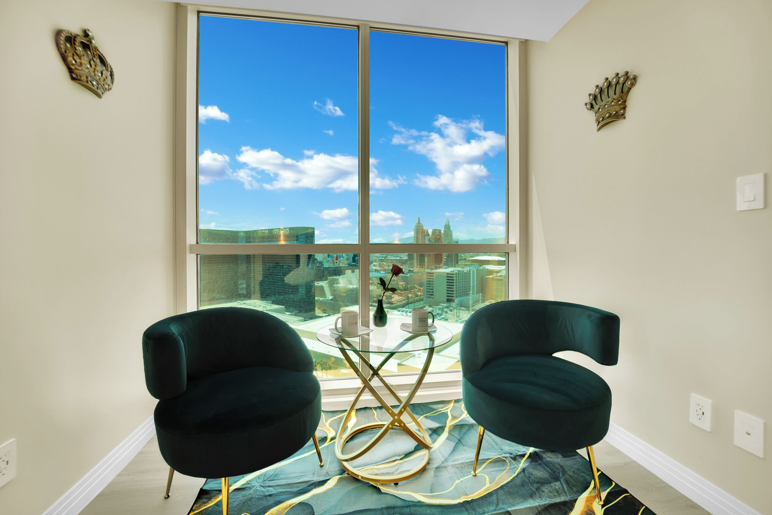 Une pièce avec deux fauteuils vert foncé, une table en verre avec un pied doré, et une vue sur le paysage urbain à travers de grandes fenêtres.