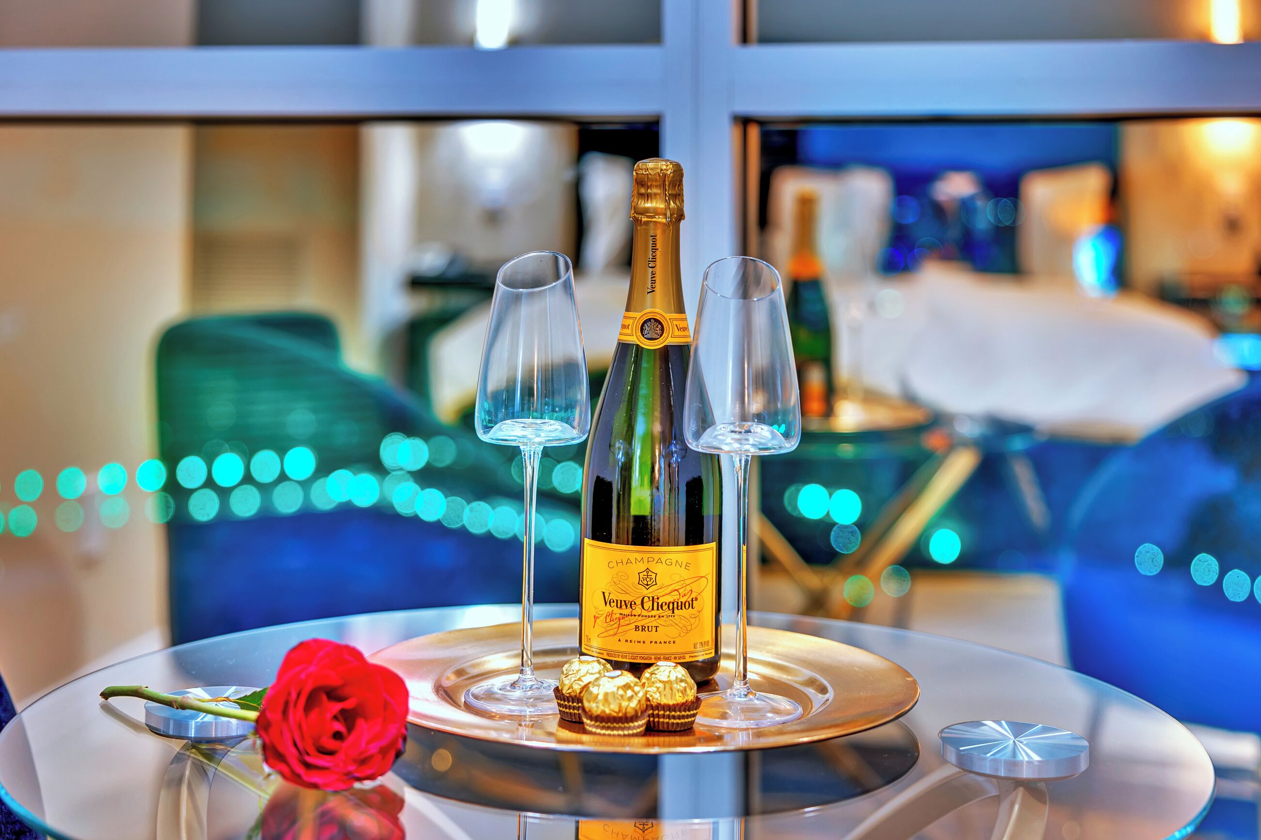 A bottle of Veuve Clicquot champagne, two glasses, a red rose, and three chocolates on a reflective surface.