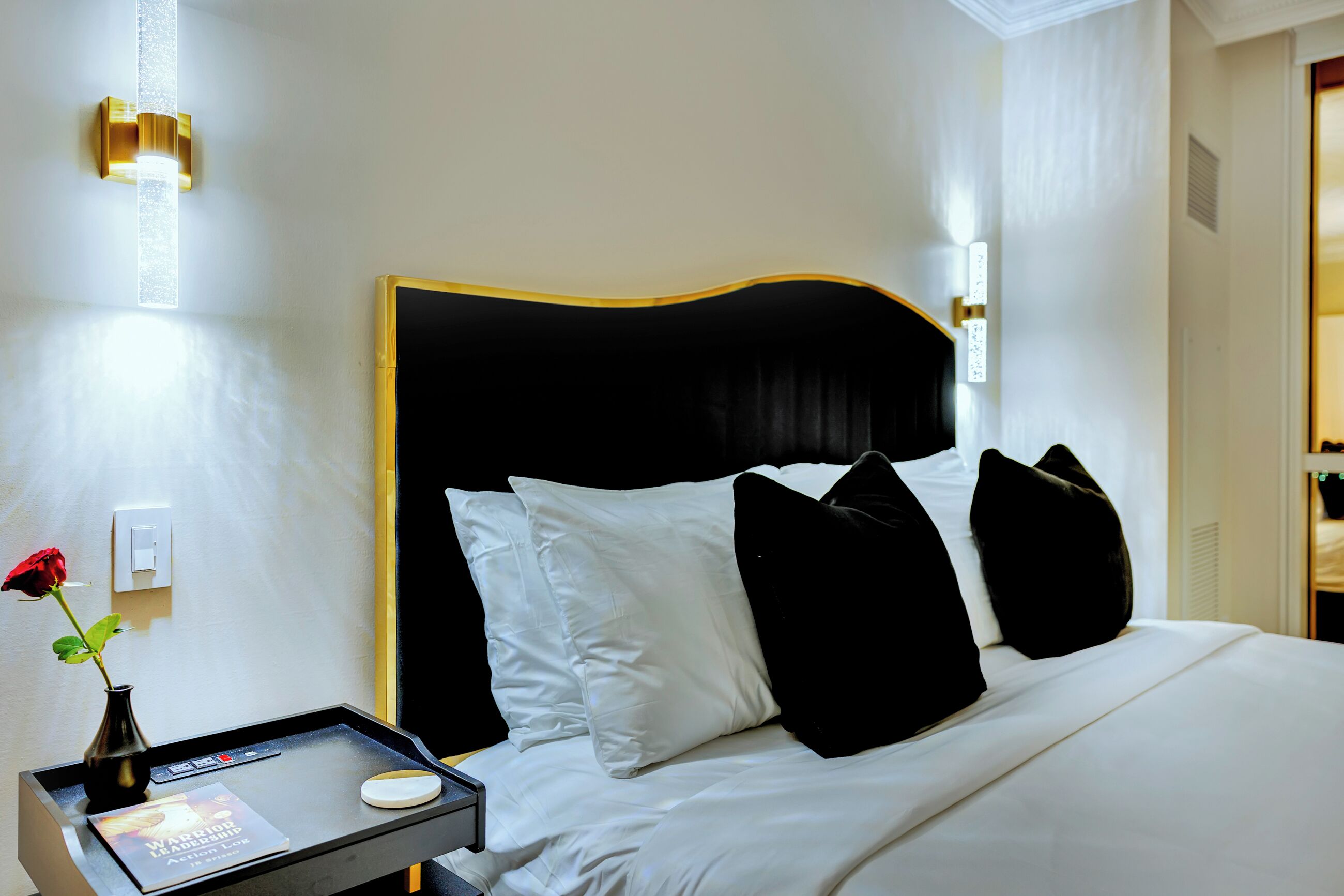 A hotel room with a bed, a bedside table with a tray, a book, and a vase with a rose.