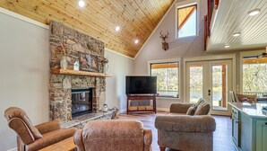 Un salón comedor amplio con chimenea de piedra, asientos cómodos, televisor y grandes ventanas con vistas a un espacio exterior pintoresco.