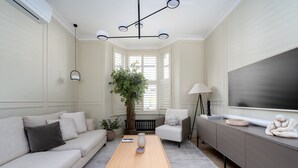 Uma sala de estar moderna com um sofá bege, uma mesa de centro de madeira, um abajur, uma televisão grande e um vaso com planta.