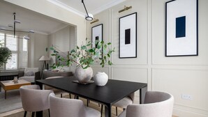 Uma sala de jantar com mesa escura, cadeiras claras e um vaso com plantas verdes.