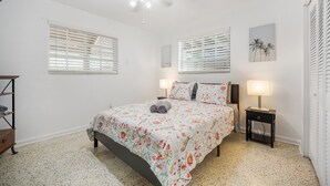 Une chambre à coucher comprenant un lit, deux tables de chevet, un ventilateur de plafond et une fenêtre avec des stores.