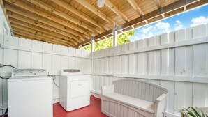 Un espace extérieur abrité, aménagé en buanderie, avec un sol rouge, un lave-linge, un sèche-linge et un banc.