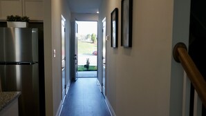 A narrow hallway with a stainless steel refrigerator on the left, a wooden handrail on the right, and a door leading to an outdoor area with a lawn and a parked car.