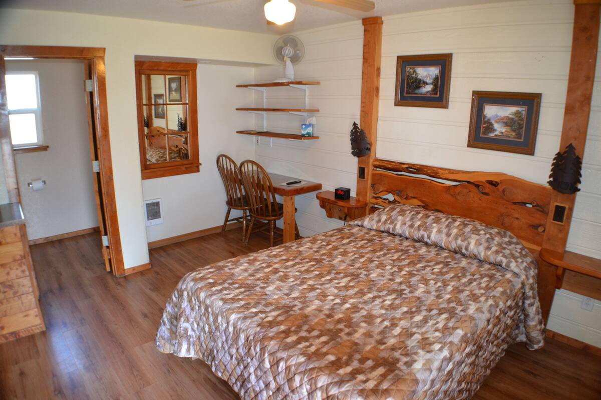 A bedroom with a wooden bed, a desk with a chair, a mirror, and shelves with framed pictures.