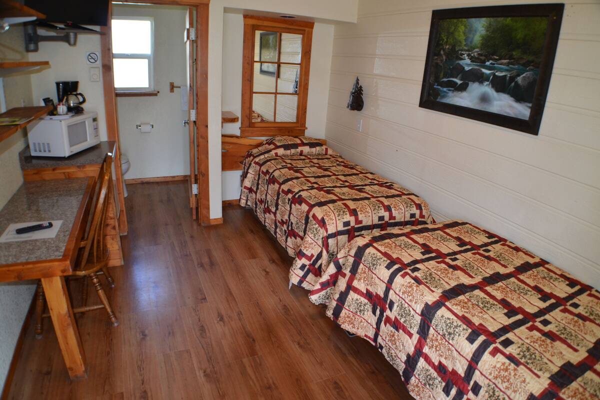 A bedroom with two beds, wooden flooring, a window, a painting, and a kitchenette area.