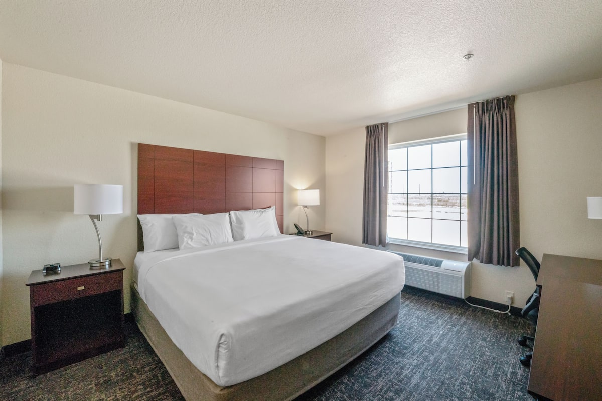 A hotel room with a large bed, a desk, a chair, a window with curtains, and a nightstand with a lamp.