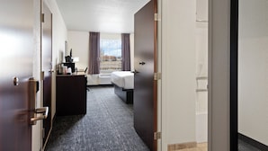 Ein Hotelzimmer mit einem Bett, einem Schreibtisch mit Lampe, einem Fenster mit Vorhängen und Blick nach draußen.