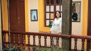 A person standing on a balcony with a wooden railing, a brown door, and a yellow wall with a window and a framed picture. - Hotel Nuestra Casa Riobamba  (Riobamba)