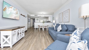 A modern living room with a blue sofa, a white console table, and a dining area with a white table and chairs. - Spacious 3 Bedroom Oceanview Condo at Sand Dunes Resort (Myrtle Beach)