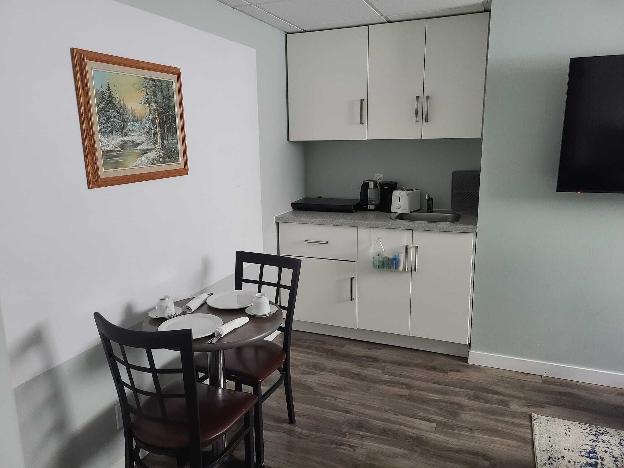Una cucina piccola e moderna con mobili bianchi, un tavolo da pranzo con sedie, un quadro appeso al muro e un televisore a schermo piatto montato sulla parete.