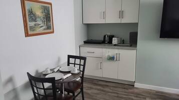 Una cucina piccola e moderna con mobili bianchi, un tavolo da pranzo con sedie, un quadro appeso al muro e un televisore a schermo piatto montato sulla parete.