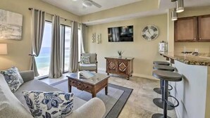 A living room with a sofa, coffee table, and a view of the ocean. There is a bar area with stools and a television mounted on the wall. - Costal Condo - Ocean & Pool Views - Sleeps 6 (Panama City Beach)