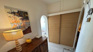 Une pièce comprenant une commode en bois, une lampe, un tableau, une armoire et une chaise recouverte d’une nappe colorée.
