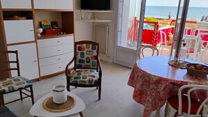 Une pièce avec une télévision, un fauteuil agrémenté de coussins colorés, une table à manger recouverte d’une nappe rouge et une vue sur la plage.