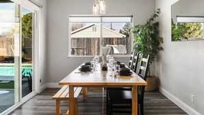 A dining area with a wooden table set for four, a large window with a view of a house and pool, and a hanging light fixture. - Cal King Hi-tech Vaca Pool Home | Ideal 4 Families (Chico)