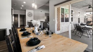 A dining area with a long wooden table set for a meal, a clock on the wall, and a view of the outside through a glass door. - Cal King Hi-tech Vaca Pool Home | Ideal 4 Families (Chico)