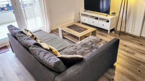 Een moderne woonkamer met een grijze bank, een houten salontafel, een tv op een witte dressoir en een vloerlamp.
