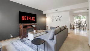 A modern living room with a grey sofa, a coffee table, a TV mounted on the wall, and a dining area in the background.