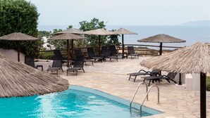 Zona de piscina con sillones de descanso y sombrillas de paja, con vistas a una masa de agua.