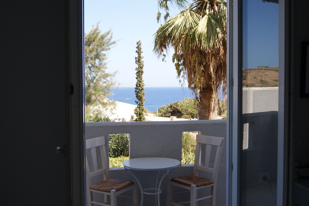 Un balcón con una mesa blanca y dos sillas, con vista al mar y palmeras a la orilla.