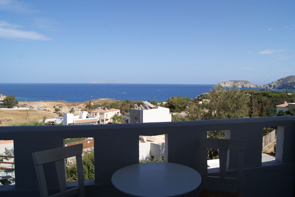 A balcony with a white table and two chairs, overlooking a coastal town with buildings and the sea.