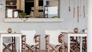 Un comedor con sillas y mesas de madera, una ventana con estantes que exhiben platos y vasos, y una decoración colgante de bambú.